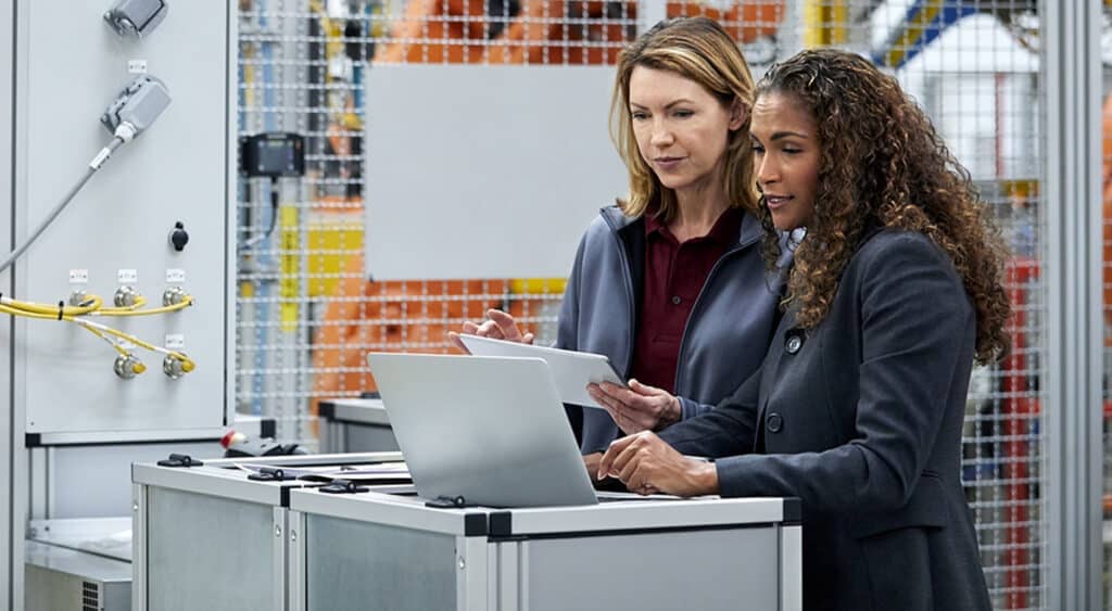 Dos mujeres vestidas profesionalmente en la fábrica revisando una tableta y una computadora portátil