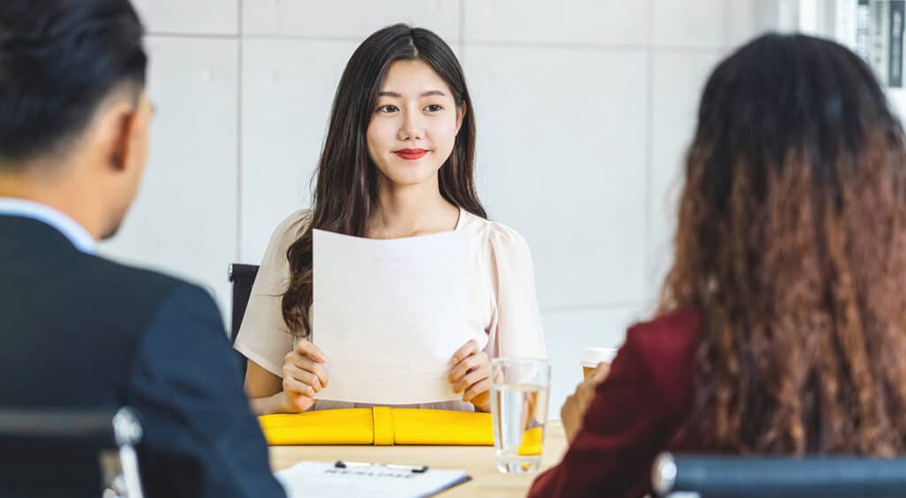 Photo of young woman holding resume ready to discuss in front of two professionals