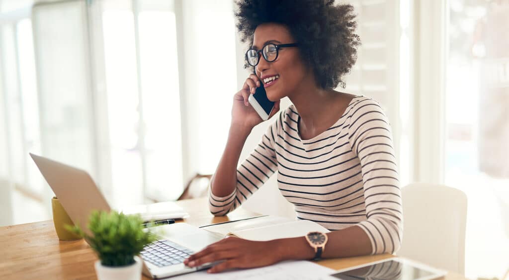 Woman in bright home office talking on cell phone with laptop open