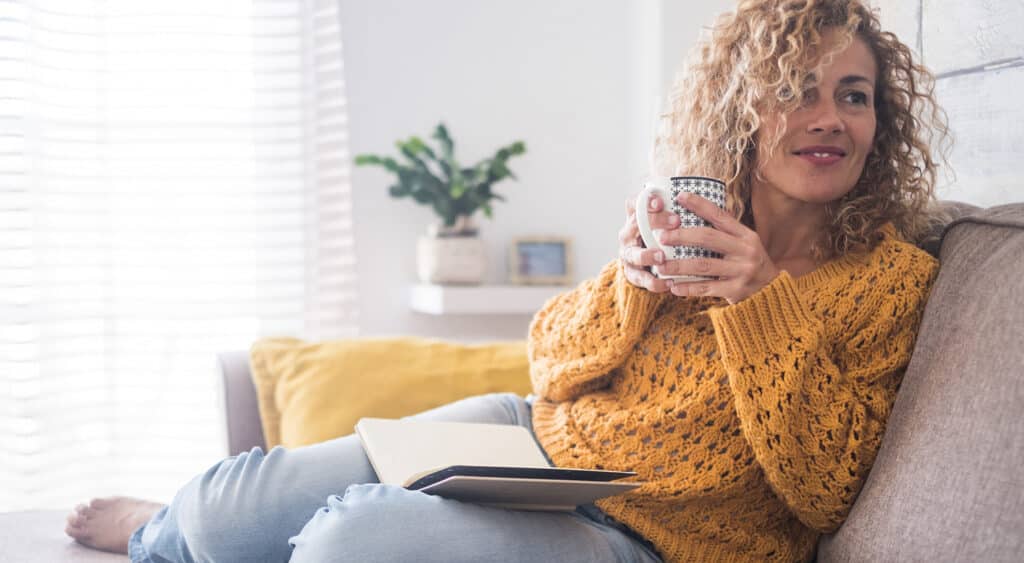 Woman sitting on couch smiling with cup of coffee and notepad