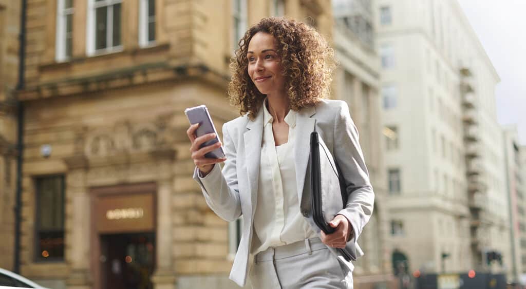 Woman smiling, holding cell phone and portfolio walking down a city street