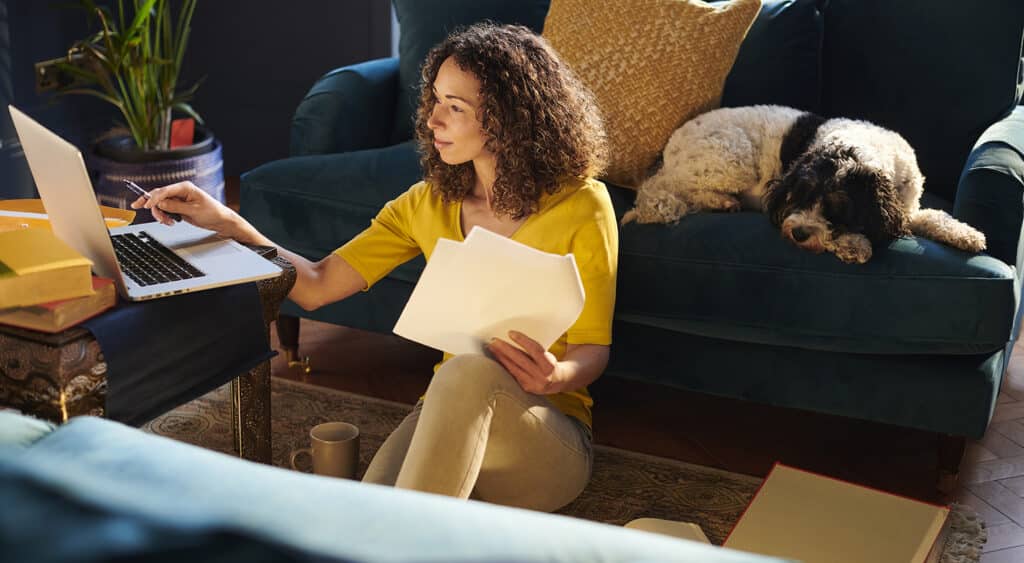 Woman working remote sitting on floor in front of laptop with dog laying on couch behind her