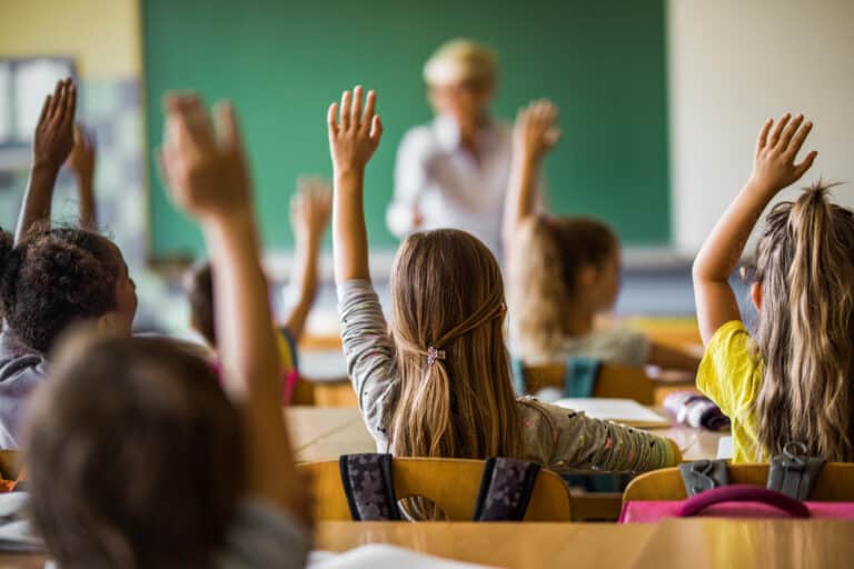 Vista trasera de estudiantes de primaria levantando los brazos en una clase.